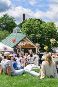 A bright, high-angle shot of a large crowd gathered on the green lawn of Seminary Park during the Indiana Peony Festival. In the foreground, a man in a grey t-shirt and baseball cap and a woman in a light-colored blouse sit on the grass, looking toward the center of the event. The park's green-roofed gazebo is visible in the middle ground, decorated with colorful flower arrangements. White event tents and more festival-goers fill the space under a blue sky with fluffy white clouds, with a tall brick chimney stack rising behind a line of lush green trees in the background.
