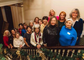 A group of approximately fifteen women are smiling and posing together on a grand, indoor staircase. They are dressed in a variety of business-casual and festive attire, including sweaters, blazers, and a sequined top. The staircase railing in the foreground is decorated with lush green holiday garlands and gold bows. The lighting is warm, and the overall atmosphere is festive and communal.