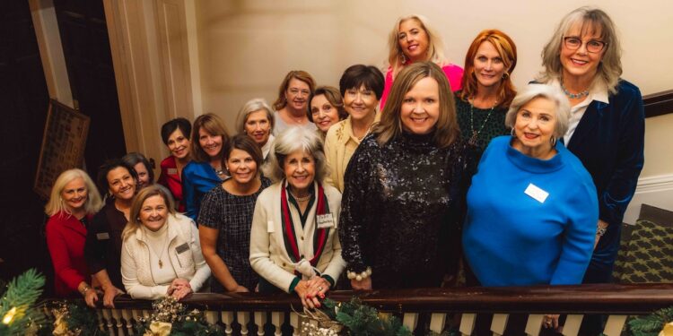 A group of approximately fifteen women are smiling and posing together on a grand, indoor staircase. They are dressed in a variety of business-casual and festive attire, including sweaters, blazers, and a sequined top. The staircase railing in the foreground is decorated with lush green holiday garlands and gold bows. The lighting is warm, and the overall atmosphere is festive and communal.