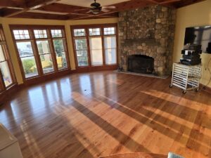 A spacious, sunlit living room featuring polished hardwood floors and a large stone fireplace with a rustic wooden mantel. Expansive windows with wooden frames wrap around the room, offering a view of a waterfront and a yellow slide outdoors. The ceiling has dark wood beams and a central ceiling fan. To the right, a television and audio equipment sit on a white rolling cart.