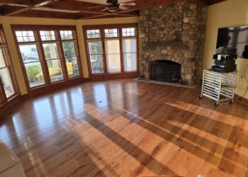 A spacious, sunlit living room featuring polished hardwood floors and a large stone fireplace with a rustic wooden mantel. Expansive windows with wooden frames wrap around the room, offering a view of a waterfront and a yellow slide outdoors. The ceiling has dark wood beams and a central ceiling fan. To the right, a television and audio equipment sit on a white rolling cart.