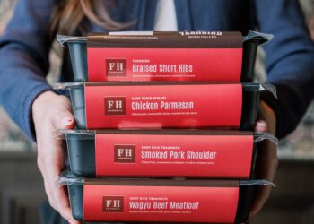 A person in a blue button-down shirt holds a stack of four black rectangular meal containers. Each container has a red label identifying the dish and the collaborating chef: Yardbird Southern Table & Bar: Braised Short Ribs (with mashed potatoes, collard greens, and sweet tea marinade). Chef Fabio Viviani: Chicken Parmesan (with penne pasta, mozzarella, and roasted tomato sauce). Chef Rick Tramonto: Smoked Pork Shoulder (with pulled pork, cornbread, and Carolina BBQ sauce). Chef Rick Tramonto: Wagyu Beef Meatloaf (with mashed potatoes, mushroom gravy, and sautéed spinach).