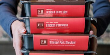 A person in a blue button-down shirt holds a stack of four black rectangular meal containers. Each container has a red label identifying the dish and the collaborating chef: Yardbird Southern Table & Bar: Braised Short Ribs (with mashed potatoes, collard greens, and sweet tea marinade). Chef Fabio Viviani: Chicken Parmesan (with penne pasta, mozzarella, and roasted tomato sauce). Chef Rick Tramonto: Smoked Pork Shoulder (with pulled pork, cornbread, and Carolina BBQ sauce). Chef Rick Tramonto: Wagyu Beef Meatloaf (with mashed potatoes, mushroom gravy, and sautéed spinach).