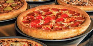 A top-down, slightly angled view of a Godfather's Pizza feast featuring five round pizzas on dark grey trays spread across a metallic surface. In the center is a golden, thick-crust pepperoni pizza with melted cheese and numerous pepperoni slices. Surrounding it are other varieties, including a pizza topped with sliced pickles and another with mixed vegetables. At the top of the frame is a dessert pizza made of small dough balls drizzled with white icing.