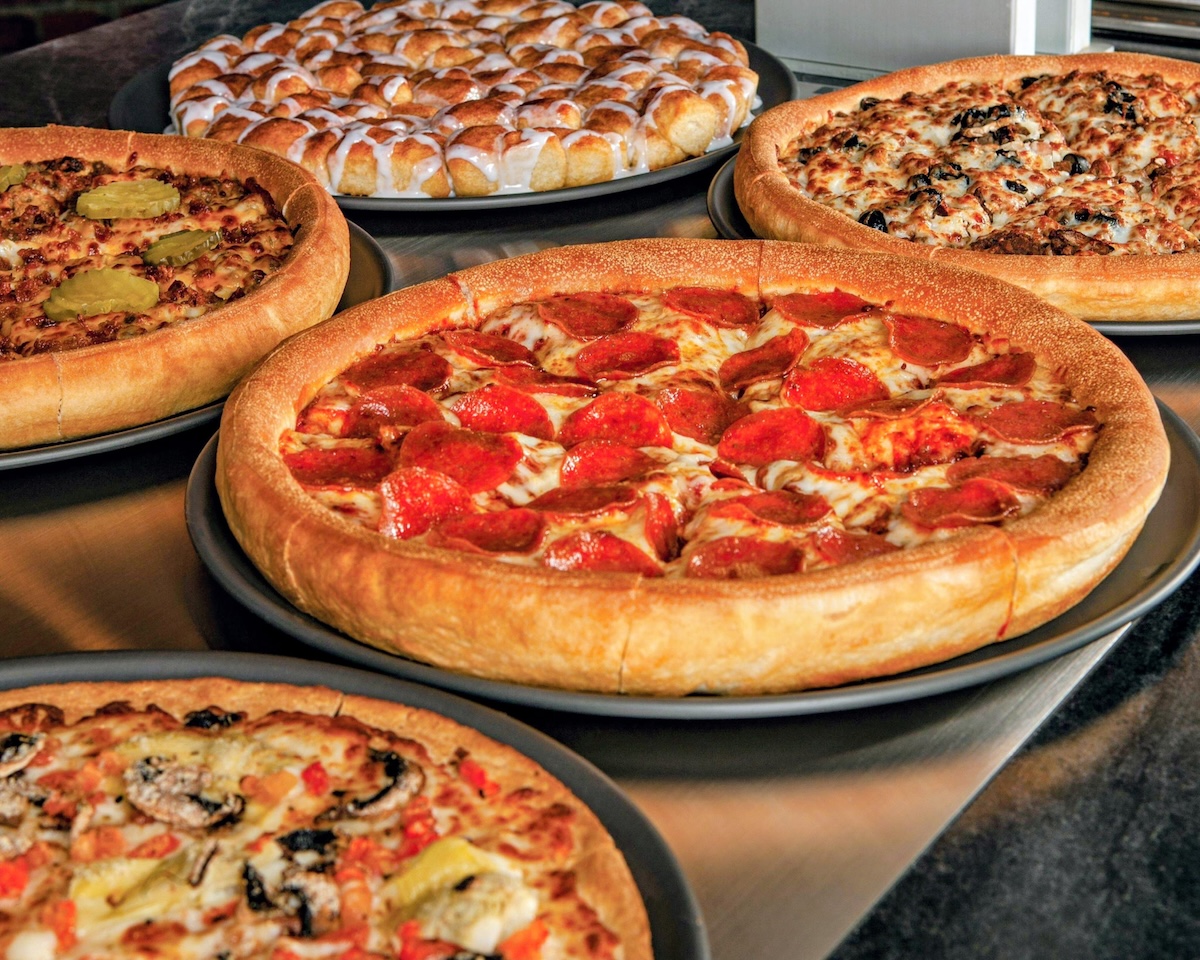 A top-down, slightly angled view of a Godfather's Pizza feast featuring five round pizzas on dark grey trays spread across a metallic surface. In the center is a golden, thick-crust pepperoni pizza with melted cheese and numerous pepperoni slices. Surrounding it are other varieties, including a pizza topped with sliced pickles and another with mixed vegetables. At the top of the frame is a dessert pizza made of small dough balls drizzled with white icing.