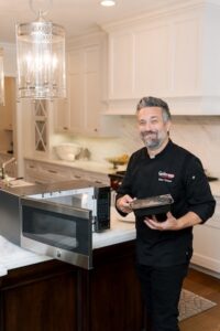 Celebrity chef Fabio Viviani stands in a bright, modern white kitchen wearing a black chef’s coat with the "FoodHaul" logo. He is smiling warmly while holding a black rectangular meal container. In the foreground, a stainless steel microwave sits open on a dark wood kitchen island with a white marble countertop.