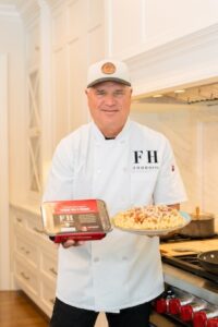 A chef, Darryl Postelnick, stands in a bright, modern white kitchen wearing a white chef’s coat with the "FH FOODHAUL" logo and a baseball cap. He is holding a packaged FoodHaul meal in his right hand and a prepared plate of the same meal—a hearty pasta dish topped with sauce and cheese—in his left. The kitchen background features white cabinetry and a high-end stainless steel stove with red knobs.