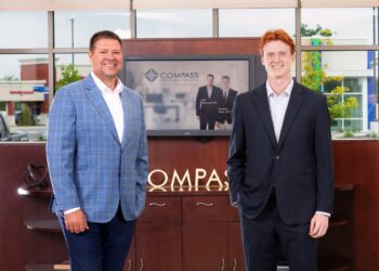 Two men in professional business attire stand smiling in a modern office lobby. On the left, a man with brown hair wears a blue windowpane patterned blazer over a white shirt and dark trousers. On the right, a younger man with red hair wears a dark navy suit with a white dress shirt. They stand in front of a dark wood cabinet featuring a silver "COMPASS" logo. A television screen in the background displays the Compass Investment Services logo and a professional photo of the two men. Large windows behind them show a bright outdoor view of a commercial area.