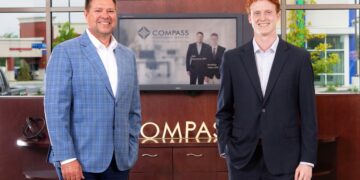 Two men in professional business attire stand smiling in a modern office lobby. On the left, a man with brown hair wears a blue windowpane patterned blazer over a white shirt and dark trousers. On the right, a younger man with red hair wears a dark navy suit with a white dress shirt. They stand in front of a dark wood cabinet featuring a silver "COMPASS" logo. A television screen in the background displays the Compass Investment Services logo and a professional photo of the two men. Large windows behind them show a bright outdoor view of a commercial area.