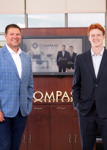 Two men in professional business attire stand smiling in a modern office lobby. On the left, a man with brown hair wears a blue windowpane patterned blazer over a white shirt and dark trousers. On the right, a younger man with red hair wears a dark navy suit with a white dress shirt. They stand in front of a dark wood cabinet featuring a silver "COMPASS" logo. A television screen in the background displays the Compass Investment Services logo and a professional photo of the two men. Large windows behind them show a bright outdoor view of a commercial area.