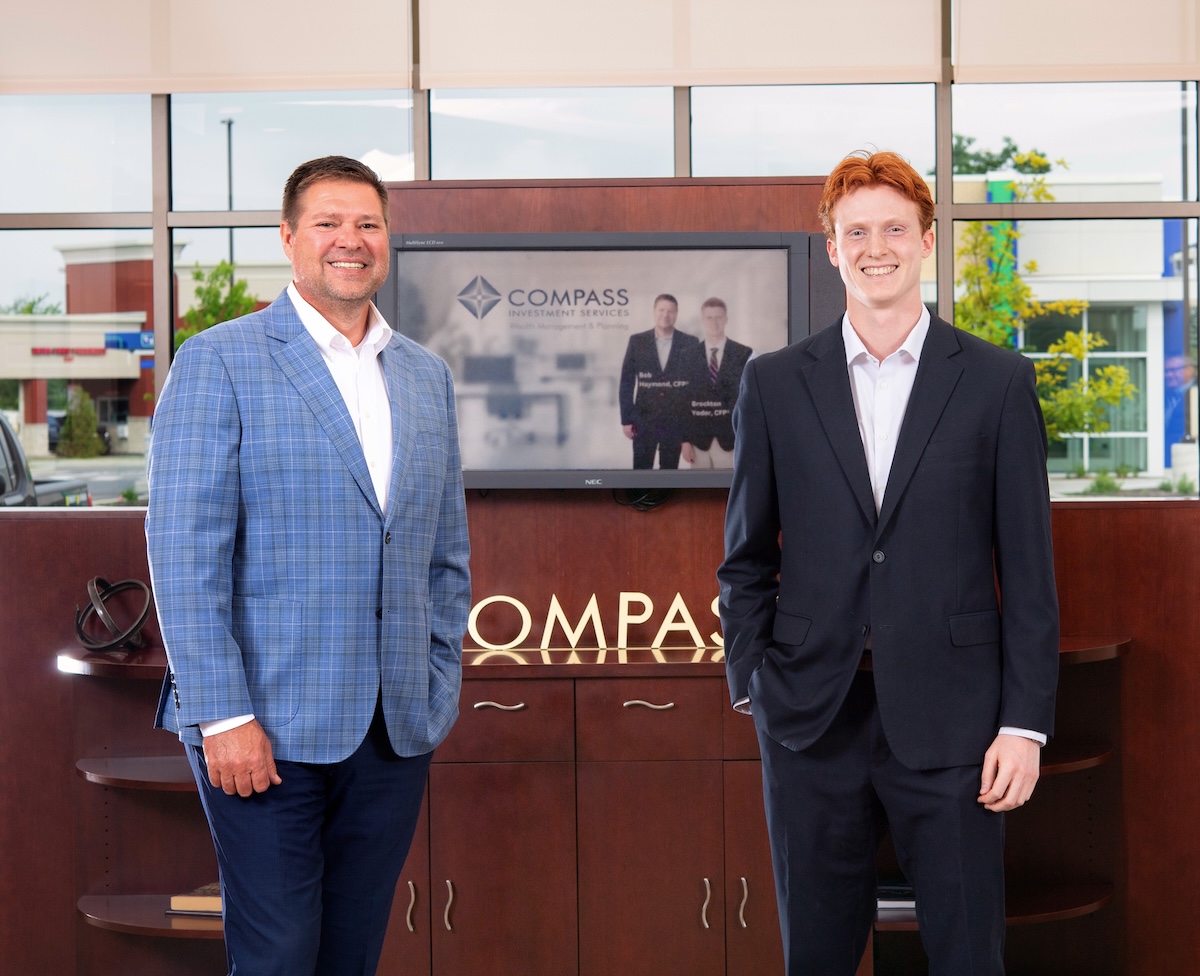 Two men in professional business attire stand smiling in a modern office lobby. On the left, a man with brown hair wears a blue windowpane patterned blazer over a white shirt and dark trousers. On the right, a younger man with red hair wears a dark navy suit with a white dress shirt. They stand in front of a dark wood cabinet featuring a silver "COMPASS" logo. A television screen in the background displays the Compass Investment Services logo and a professional photo of the two men. Large windows behind them show a bright outdoor view of a commercial area.