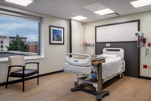 A bright and modern private hospital room featuring a single bed with white linens and a grey headboard. A light-colored wood-patterned floor covers the space, and a large window to the left offers a view of a nearby building under a cloudy sky. To the left of the bed is a tan and black armchair, and a mobile bedside table sits next to the bed. The room is well-lit by several recessed square ceiling lights and features a framed piece of artwork on the wall.