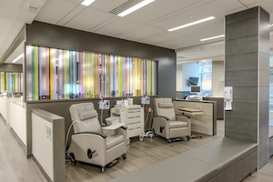 A bright and modern medical infusion center featuring two comfortable, tan-colored reclining chairs separated by a white mobile medical cart. The room has light wood-look flooring and a distinctive architectural wall with vertical glass panels in a spectrum of colors like yellow, purple, and green. A grey, floor-to-ceiling column and a built-in grey bench seat are in the foreground. The space is well-lit with recessed ceiling lights and a clean, professional aesthetic.