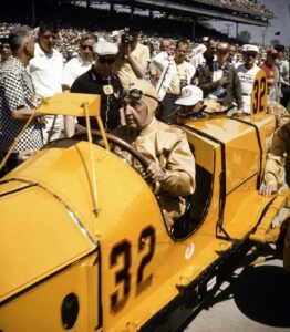 A historic photograph capturing Ray Harroun seated in the iconic yellow Marmon Wasp (No. 32) during the 1961 Golden Anniversary of the Indianapolis 500. Harroun, wearing a tan racing suit and a leather aviator-style cap with goggles, is surrounded by a crowd of spectators and photographers at the Indianapolis Motor Speedway. The car’s distinctive pointed cowl and the large black number "32" on its side are prominently displayed.