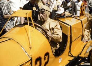 A historic photograph capturing Ray Harroun seated in the iconic yellow Marmon Wasp (No. 32) during the 1961 Golden Anniversary of the Indianapolis 500. Harroun, wearing a tan racing suit and a leather aviator-style cap with goggles, is surrounded by a crowd of spectators and photographers at the Indianapolis Motor Speedway. The car’s distinctive pointed cowl and the large black number "32" on its side are prominently displayed.