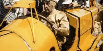 A historic photograph capturing Ray Harroun seated in the iconic yellow Marmon Wasp (No. 32) during the 1961 Golden Anniversary of the Indianapolis 500. Harroun, wearing a tan racing suit and a leather aviator-style cap with goggles, is surrounded by a crowd of spectators and photographers at the Indianapolis Motor Speedway. The car’s distinctive pointed cowl and the large black number "32" on its side are prominently displayed.