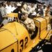 A historic photograph capturing Ray Harroun seated in the iconic yellow Marmon Wasp (No. 32) during the 1961 Golden Anniversary of the Indianapolis 500. Harroun, wearing a tan racing suit and a leather aviator-style cap with goggles, is surrounded by a crowd of spectators and photographers at the Indianapolis Motor Speedway. The car’s distinctive pointed cowl and the large black number "32" on its side are prominently displayed.