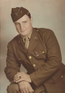 A sepia-toned vintage studio portrait of a young man in a mid-20th-century U.S. Army uniform. He is wearing a dark Garrison cap with a small insignia, a collared shirt with a necktie, and a four-pocket service coat. On his lapels are circular "U.S." and branch-of-service disks, and a marksmanship badge is pinned to his left breast pocket. He is leaning forward with his hands crossed, revealing a ring on his finger, and looking toward the camera with a slight, pleasant smile.