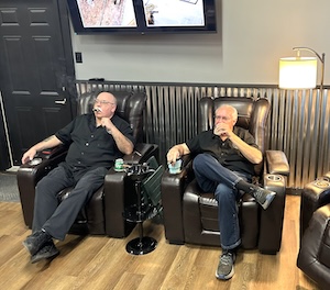 Two men relax in large, black leather recliners inside a lounge. The man on the left, who is bald and wears glasses and a black button-down shirt, holds a cigar and looks toward a large TV screen mounted on the wall. The man on the right has short white hair and a beard, is also dressed in a black shirt, and holds a drink. The room features light wood-look flooring, a warm lamp on the right, and a distinctive wall with a corrugated metal wainscoting.