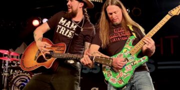 Two guitarists perform together on a dimly lit stage with red and white stage lights in the background. On the left, wears a wide-brimmed tan hat with a large feather, a black "MAMA TRIED" t-shirt, and has his beard styled in long braids. He is playing an acoustic Taylor guitar that features a distinctive red sticker reading "FRIENDS DON'T LET FRIENDS GET FRIENDS HAIRCUTS..."—a famous tribute to Alice in Chains. On the right, has long straight hair and wears a black "BAD MOTHER..." t-shirt. He is playing a vibrant green Kramer electric guitar completely covered in various colorful stickers.