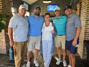 Four men and one woman are standing together and smiling in a well-lit indoor space with brick walls and a dark tiled floor. The woman in the center is wearing blue and white striped pants and holding a white envelope. The men are dressed in casual golf attire, including polo shirts and baseball caps. A television and a green plant are visible in the background, suggesting a clubhouse or restaurant setting.