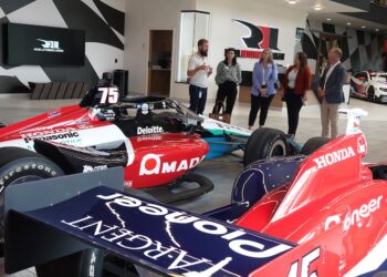 Five visitors listening to a guide in the RLL Racing showroom in Zionsville. In the foreground is a No. 75 IndyCar with prominent AMADA and Honda branding.