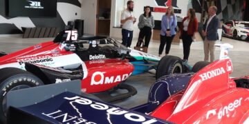 Five visitors listening to a guide in the RLL Racing showroom in Zionsville. In the foreground is a No. 75 IndyCar with prominent AMADA and Honda branding.