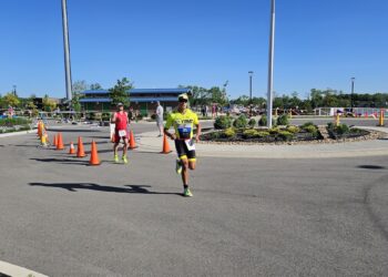 A sunny outdoor scene at the Geist Waterfront Triathlon showing participants running on an asphalt course through a parking area with a blue-roofed building and Geist Reservoir in the background.