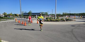 A sunny outdoor scene at the Geist Waterfront Triathlon showing participants running on an asphalt course through a parking area with a blue-roofed building and Geist Reservoir in the background.