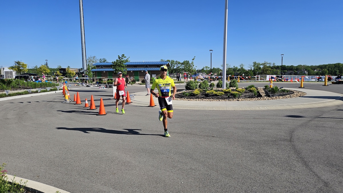 A sunny outdoor scene at the Geist Waterfront Triathlon showing participants running on an asphalt course through a parking area with a blue-roofed building and Geist Reservoir in the background.
