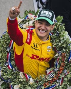 A close-up portrait of Álex Palou, the winner of the 109th Indy 500, smiling joyfully in Victory Circle. He is wearing a bright yellow and red Chip Ganassi Racing firesuit featuring the DHL and Honda logos. A large, lush winner's wreath, decorated with white orchids and checkered flags and featuring the "BORGWARNER" name, is draped over his shoulders. He wears a green and white baseball cap that reads "Indy 500 109th Running" and is pointing one finger toward the sky in a celebratory gesture.