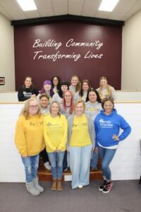 A diverse group of over a dozen women stand together for a group photo in an office or community center. They are posed on and around a small wooden staircase in front of a deep maroon wall that reads "Building Community, Transforming Lives" in white cursive script. Many are wearing branded "Family Promise" hoodies in vibrant blue, bright yellow, and black. They are all smiling warmly at the camera.