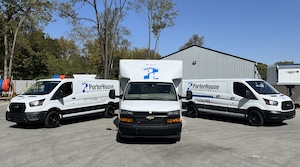 Three white commercial service vehicles from PorterHouse Contracting LLC are parked in a semi-circle on a gray paved lot. The fleet consists of two Ford Transit vans on the sides and one larger Chevrolet box truck in the center. Each vehicle is branded with a blue and gray "P" logo and the company name, "PorterHouse," on the sides. The background features a gray building, mature green trees under a clear blue sky, and other construction-related trailers partially visible.