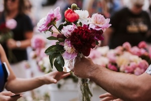 A close-up, shallow-focus shot at the Indiana Peony Festival showing a hand holding a lush, hand-tied bouquet of fresh peonies. The bouquet features a mix of vibrant blooms, including deep burgundy, bright red, and soft pink peonies in various stages of opening. In the blurred background, another hand reaches toward the flowers, and several other colorful peony arrangements are visible on a table, capturing the bustling atmosphere of the festival in Seminary Park.