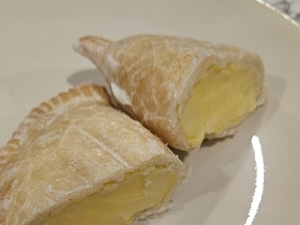 A flaky, golden-brown hand pie is cut in half on a cream-colored plate, showcasing a generous, smooth lemon custard filling. The pastry features a traditional crimped edge and a lightly glazed, shattered-look crust. The creamy yellow filling is thick and holds its shape, indicating a rich and zesty dessert.