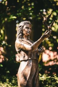 A bronze sculpture of a woman with long hair, wearing a simple sleeveless dress. She is looking slightly upward with a peaceful expression, her right arm extended as if releasing two small birds into flight. The sculpture is situated outdoors, surrounded by soft-focus green foliage and dappled sunlight, creating a serene and natural atmosphere.