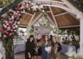 Four smiling young women stand under a large wooden gazebo at Seminary Park during the Indiana Peony Festival. The gazebo's white-painted entrance is draped with a vibrant floral arch of pink, red, and white peonies. The women are dressed in casual attire; two hold hand-tied flower bouquets. A "Thank You to Our Sponsors" banner is visible in the background, featuring logos from local supporters like Smith’s Jewelers and CCHA Law. The setting is bright and lively, with other festival attendees and white-clothed tables blurred in the background.