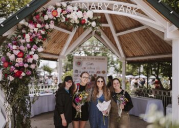 Four smiling young women stand under a large wooden gazebo at Seminary Park during the Indiana Peony Festival. The gazebo's white-painted entrance is draped with a vibrant floral arch of pink, red, and white peonies. The women are dressed in casual attire; two hold hand-tied flower bouquets. A "Thank You to Our Sponsors" banner is visible in the background, featuring logos from local supporters like Smith’s Jewelers and CCHA Law. The setting is bright and lively, with other festival attendees and white-clothed tables blurred in the background.