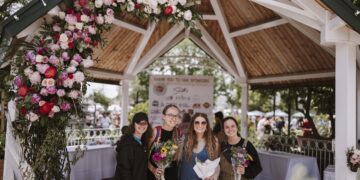 Four smiling young women stand under a large wooden gazebo at Seminary Park during the Indiana Peony Festival. The gazebo's white-painted entrance is draped with a vibrant floral arch of pink, red, and white peonies. The women are dressed in casual attire; two hold hand-tied flower bouquets. A "Thank You to Our Sponsors" banner is visible in the background, featuring logos from local supporters like Smith’s Jewelers and CCHA Law. The setting is bright and lively, with other festival attendees and white-clothed tables blurred in the background.