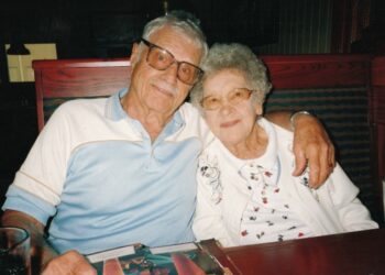 heartwarming, candid photo of an elderly couple sitting together in a restaurant booth. The man, on the left, has grey hair and a mustache, wearing large glasses and a light blue and white polo shirt. He has his arm affectionately around the woman, who has short, curly white hair and glasses, and is wearing a white cardigan with delicate floral embroidery. They are both smiling gently at the camera, with a menu and water glass visible on the table in front of them.