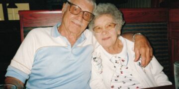 heartwarming, candid photo of an elderly couple sitting together in a restaurant booth. The man, on the left, has grey hair and a mustache, wearing large glasses and a light blue and white polo shirt. He has his arm affectionately around the woman, who has short, curly white hair and glasses, and is wearing a white cardigan with delicate floral embroidery. They are both smiling gently at the camera, with a menu and water glass visible on the table in front of them.