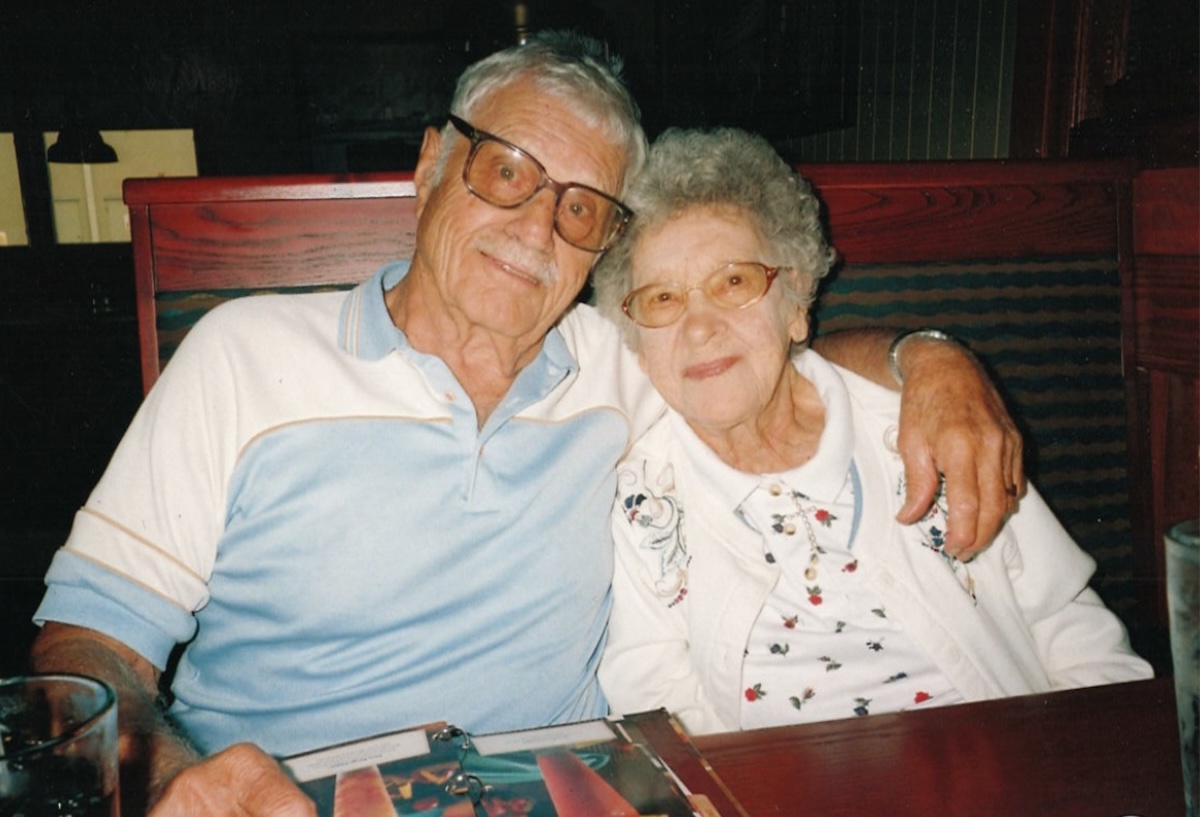 heartwarming, candid photo of an elderly couple sitting together in a restaurant booth. The man, on the left, has grey hair and a mustache, wearing large glasses and a light blue and white polo shirt. He has his arm affectionately around the woman, who has short, curly white hair and glasses, and is wearing a white cardigan with delicate floral embroidery. They are both smiling gently at the camera, with a menu and water glass visible on the table in front of them.