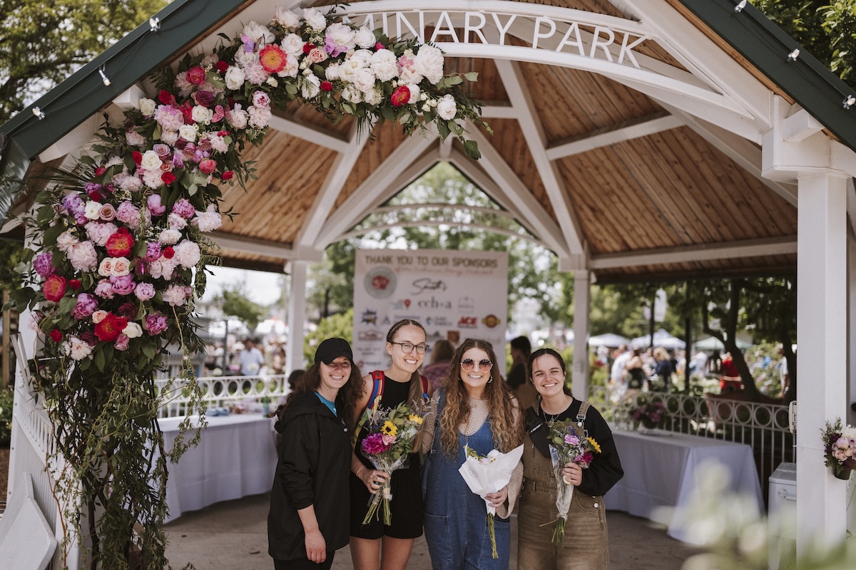 Four smiling young women stand under a large wooden gazebo at Seminary Park during the Indiana Peony Festival. The gazebo's white-painted entrance is draped with a vibrant floral arch of pink, red, and white peonies. The women are dressed in casual attire; two hold hand-tied flower bouquets. A "Thank You to Our Sponsors" banner is visible in the background, featuring logos from local supporters like Smith’s Jewelers and CCHA Law. The setting is bright and lively, with other festival attendees and white-clothed tables blurred in the background.