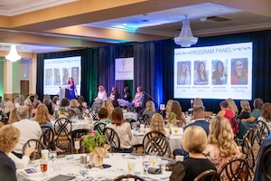 A large ballroom filled with people seated at round banquet tables during a program panel. In the background, a stage features a speaker at a podium and four panelists seated in chairs. Large projector screens on either side of the stage display the "Mentoring Program Panel" with photos and names of the participants. The room is decorated with elegant chandeliers and professional event lighting.