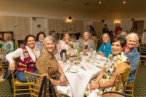 A group of eight women are smiling and gathered around a circular dining table set with a white tablecloth. They are seated in elegant gold Chiavari chairs within a well-lit banquet hall. The table is set with formal dinnerware, water glasses, and a thermal carafe. The women are dressed in colorful, casual-chic attire, including floral prints, leopard patterns, and a vibrant patchwork vest, creating a warm and social atmosphere.