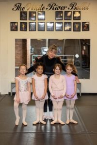 A medium full-shot of Diana Ephlin, founder of The Dancin' Place, posing with four young ballet students in her Noblesville studio. Diana stands in the back, dressed in black with her arms around the girls. The four young dancers, likely in a primary ballet class, stand in a row wearing pink and purple leotards, tights, and ballet slippers, with their hands held gracefully in front. They are positioned in front of a large studio mirror under the elegant gold script "The White River Ballet" and a gallery of framed performance photos.