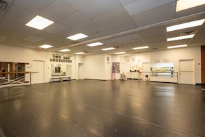 A wide-angle, low-perspective shot of a large, empty dance studio at The Dancin' Place in Noblesville, Indiana. The room features a professional black dance floor that reflects the bright, recessed square ceiling lights. White walls surround the space, with a large mirror on the left and a wall of framed performance photos and the script "The White River Ballet" in the background. Portable wooden ballet barres and two white doors are positioned along the walls. The studio is clean and well-lit, creating an open and inviting atmosphere for practice.