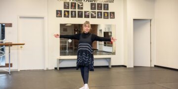 A full-shot of an older woman, Diana Ephlin, the founder of The Dancin' Place, standing in the center of her ballet studio in Noblesville. She has her arms outstretched in a welcoming gesture, wearing a black turtleneck under a black Free People trapeze slip dress with white lace chevron patterns, black leggings, and white sneakers. The studio features a black dance floor, white walls, and a large mirror. Above the mirror, the words "The White River Ballet" are written in elegant gold script, surrounded by a gallery of framed performance photos.