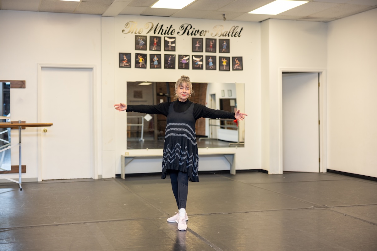 A full-shot of an older woman, Diana Ephlin, the founder of The Dancin' Place, standing in the center of her ballet studio in Noblesville. She has her arms outstretched in a welcoming gesture, wearing a black turtleneck under a black Free People trapeze slip dress with white lace chevron patterns, black leggings, and white sneakers. The studio features a black dance floor, white walls, and a large mirror. Above the mirror, the words "The White River Ballet" are written in elegant gold script, surrounded by a gallery of framed performance photos.