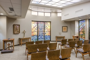 A bright, serene hospital chapel with rows of modern wooden chairs on a light-colored floor. The room features a prominent central stained-glass window with vibrant orange and amber patterns, flanked by additional narrow stained-glass windows. To the left, there is a wooden podium with a cross and a small table with a crucifix. The space is illuminated by a large grid of recessed ceiling lights and high clerestory windows that fill the room with natural light.